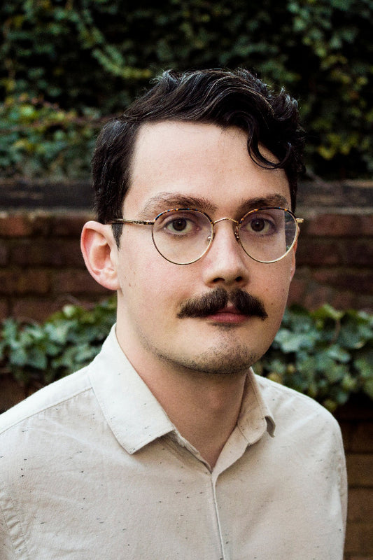 Man with glasses and a mustache wearing a light-colored shirt against a brick wall and greenery background.