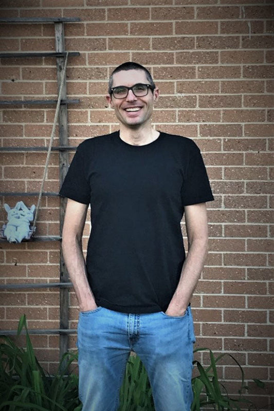 Man wearing a black t-shirt and blue jeans standing against a brick wall.